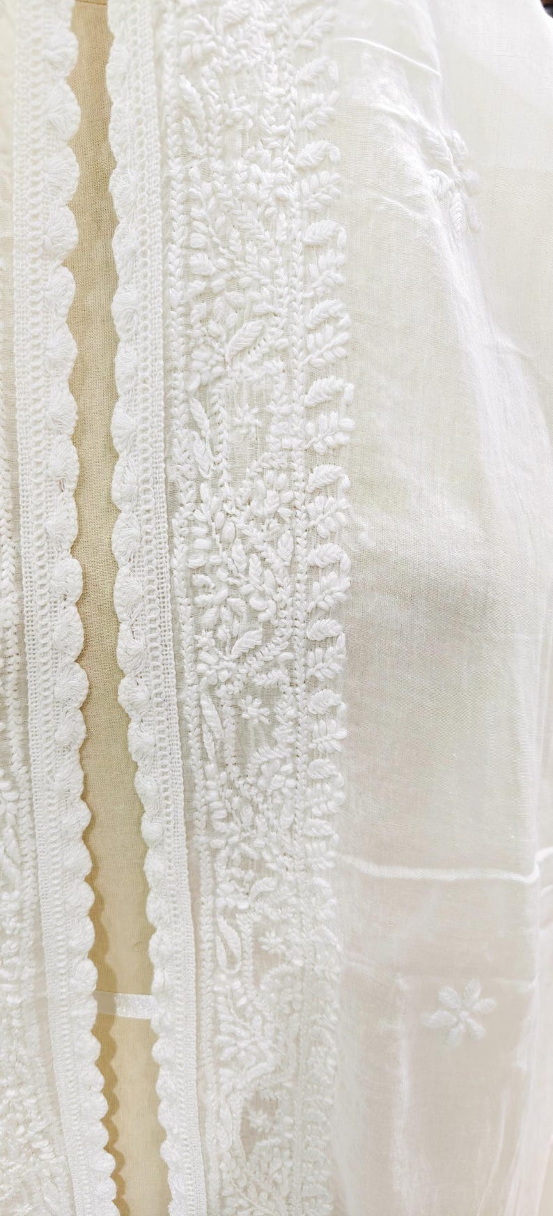 May include: Close-up of white fabric with intricate embroidery. The design features floral and leaf patterns along the edges and center. The fabric has a delicate, sheer appearance, suggesting a lightweight garment or textile.