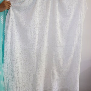 May include: White embroidered sheer fabric with a delicate floral pattern. The fabric has a scalloped edge and is draped over a hand.
