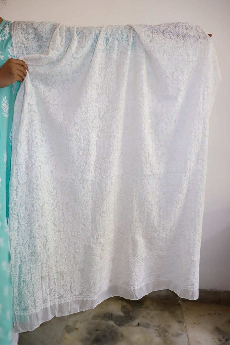 May include: White embroidered sheer fabric with a delicate floral pattern. The fabric has a scalloped edge and is draped over a hand.