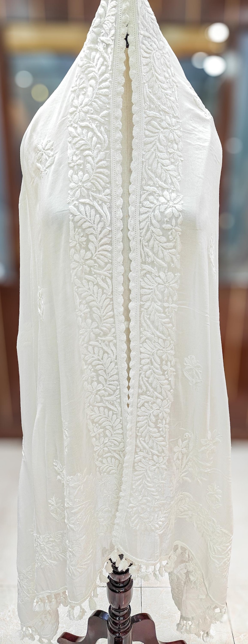 May include: White embroidered fabric with floral and leaf patterns. The fabric is draped over a form, showcasing the intricate detailing of the design. The embroidery is visible on the edges and center of the fabric.