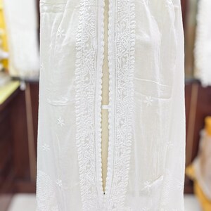 May include: A white, sheer fabric garment with intricate embroidered detailing. The garment is draped over a mannequin, showcasing the delicate floral and geometric patterns. The fabric appears lightweight and airy, suitable for a shawl or scarf.