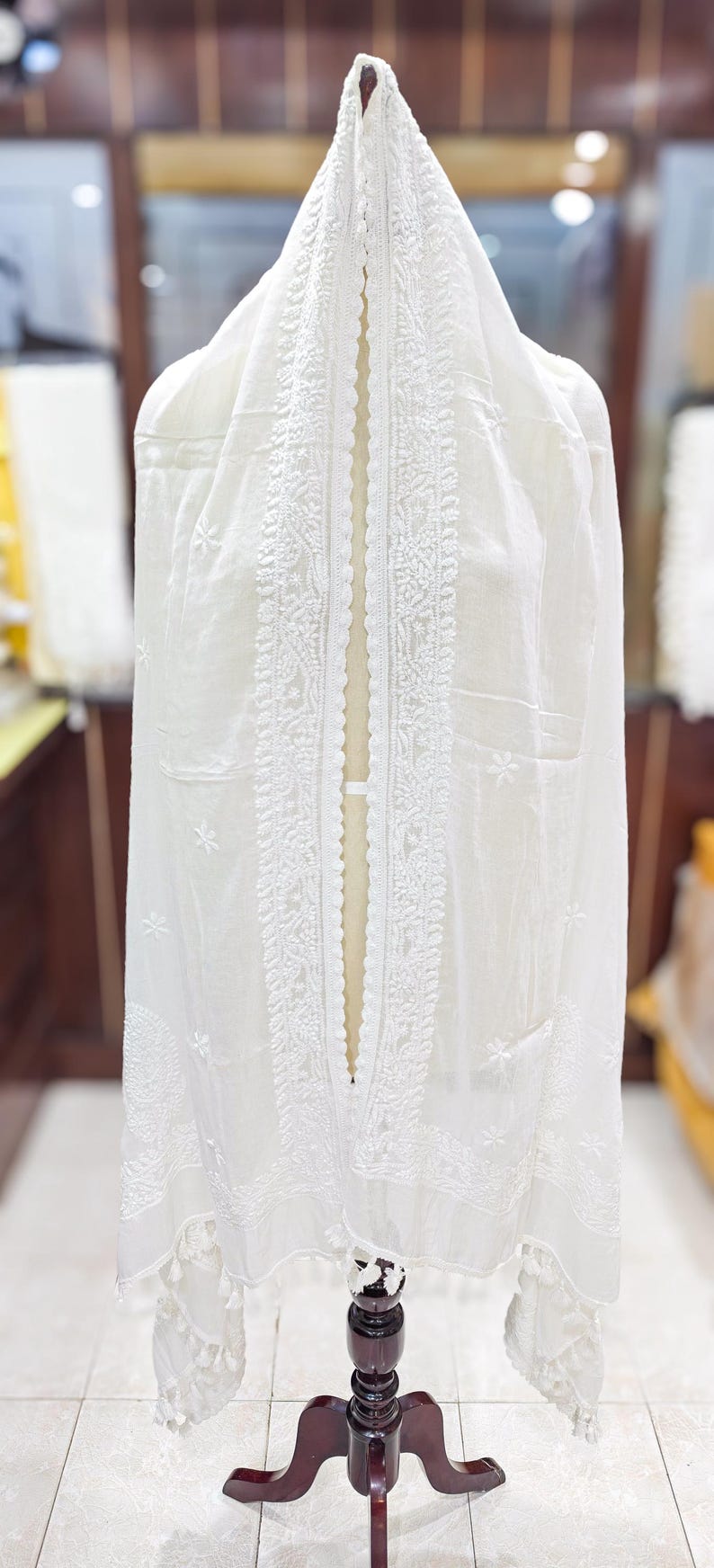 May include: A white, sheer fabric garment with intricate embroidered detailing. The garment is draped over a mannequin, showcasing the delicate floral and geometric patterns. The fabric appears lightweight and airy, suitable for a shawl or scarf.