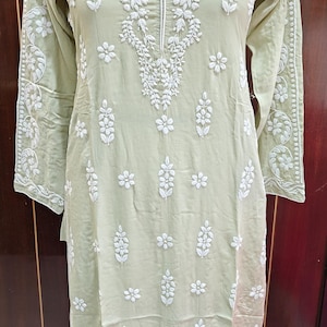 May include: A light green tunic dress with intricate white floral embroidery. The long-sleeved dress features a V-neck and detailed patterns along the sleeves and bodice. The dress is displayed on a mannequin, showcasing its flowing design.