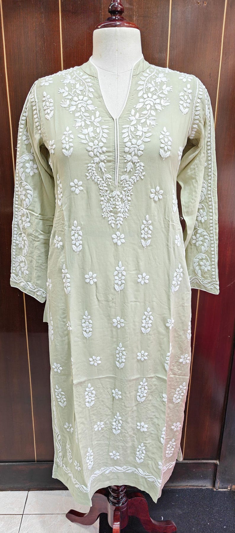 May include: A light green tunic dress with intricate white floral embroidery. The long-sleeved dress features a V-neck and detailed patterns along the sleeves and bodice. The dress is displayed on a mannequin, showcasing its flowing design.