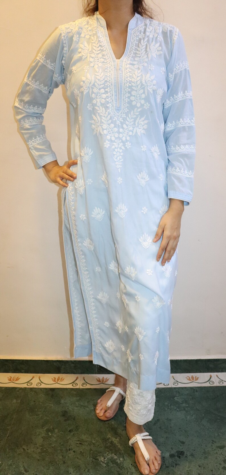 May include: A light blue embroidered kurta and white pants. The kurta features intricate white floral embroidery along the neckline, sleeves, and body. The pants are white and have a textured appearance. The outfit is worn with white sandals.