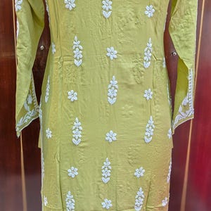 Puede incluir: Una t&uacute;nica verde claro con bordados florales blancos. La t&uacute;nica de manga larga presenta un cuello redondo y un corte recto. El bordado se concentra en la parte delantera y las mangas, con un motivo floral. La tela parece ser ligera.