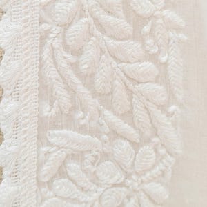 May include: Close-up of white fabric with intricate floral embroidery. The design features detailed leaves and flowers, with a textured, raised appearance. A decorative border with a woven pattern runs along the left side.