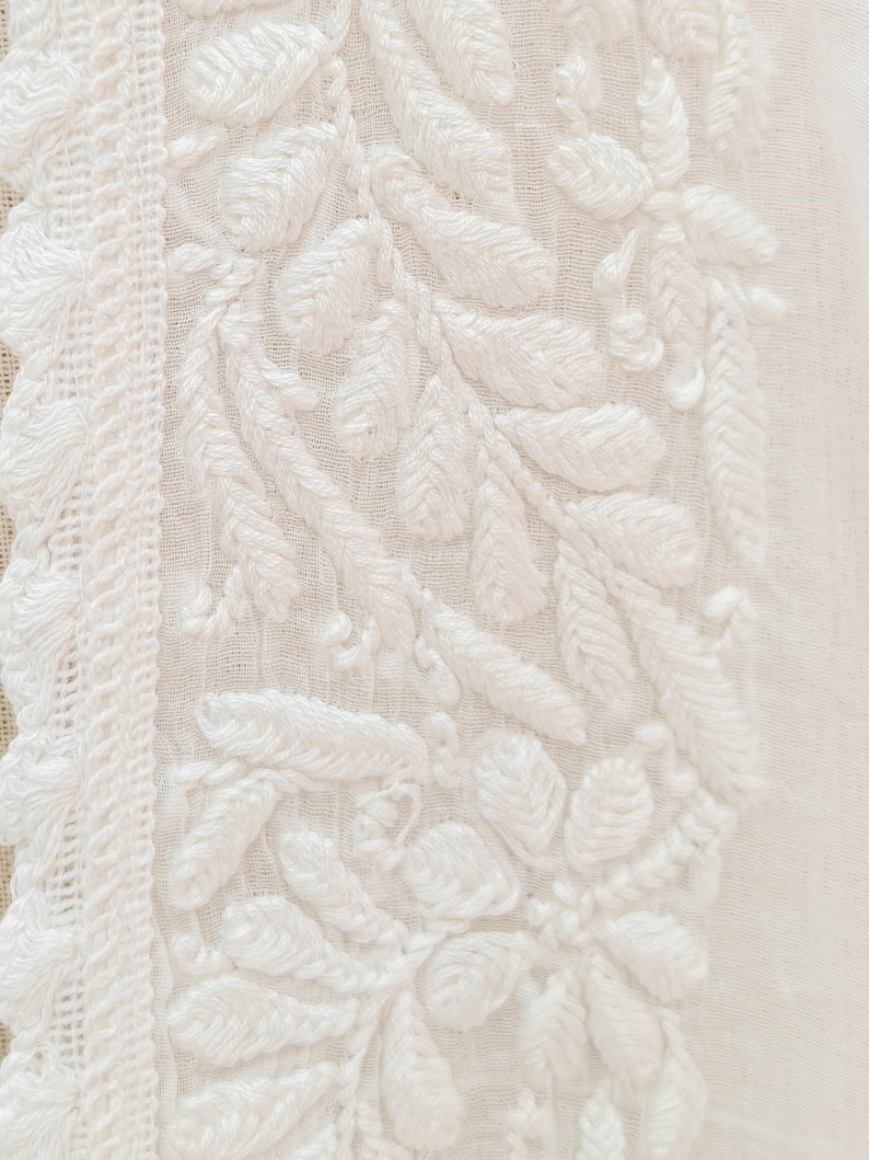 May include: Close-up of white fabric with intricate floral embroidery. The design features detailed leaves and flowers, with a textured, raised appearance. A decorative border with a woven pattern runs along the left side.
