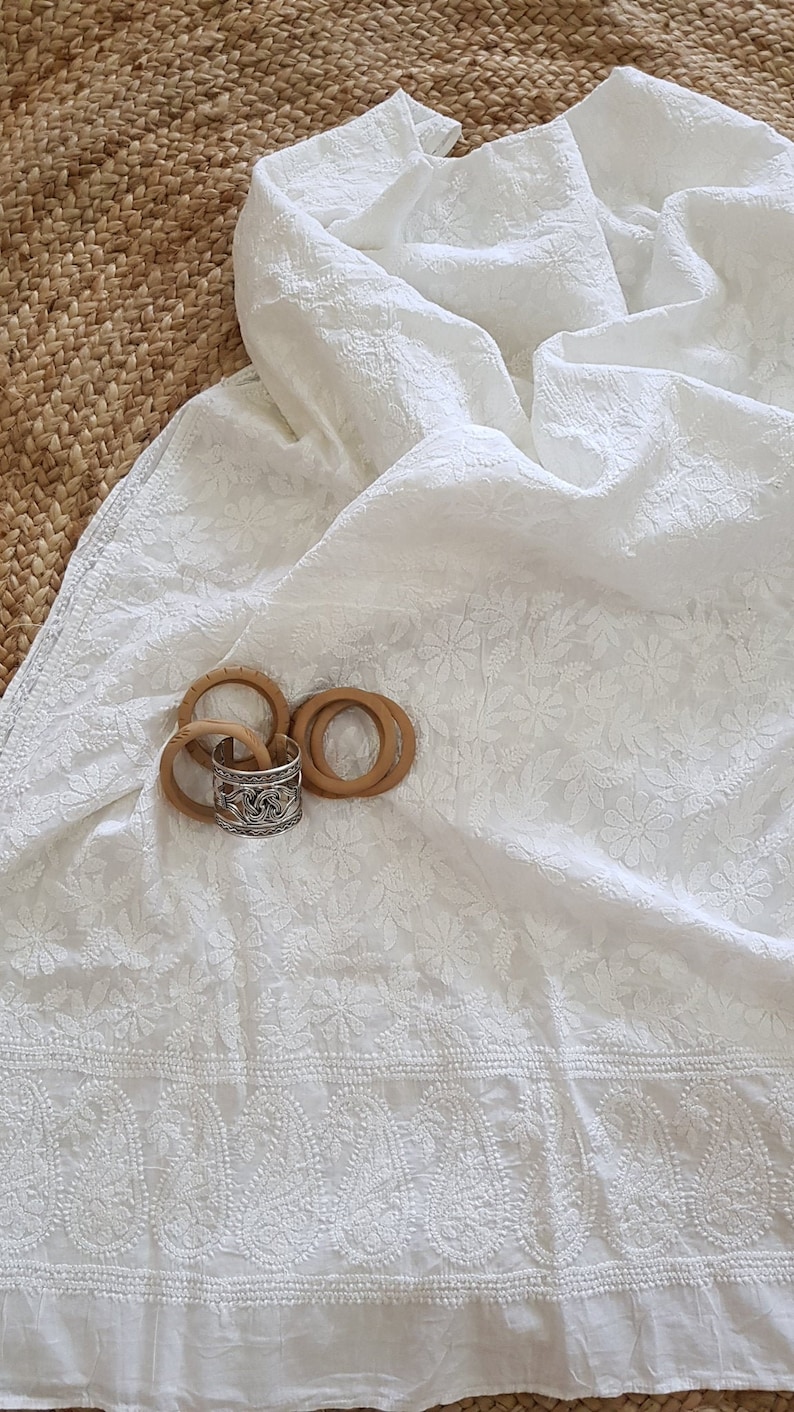 May include: White cotton fabric with delicate floral embroidery. The fabric is folded and draped over a woven surface. There are four wooden rings and a silver ring on the fabric.