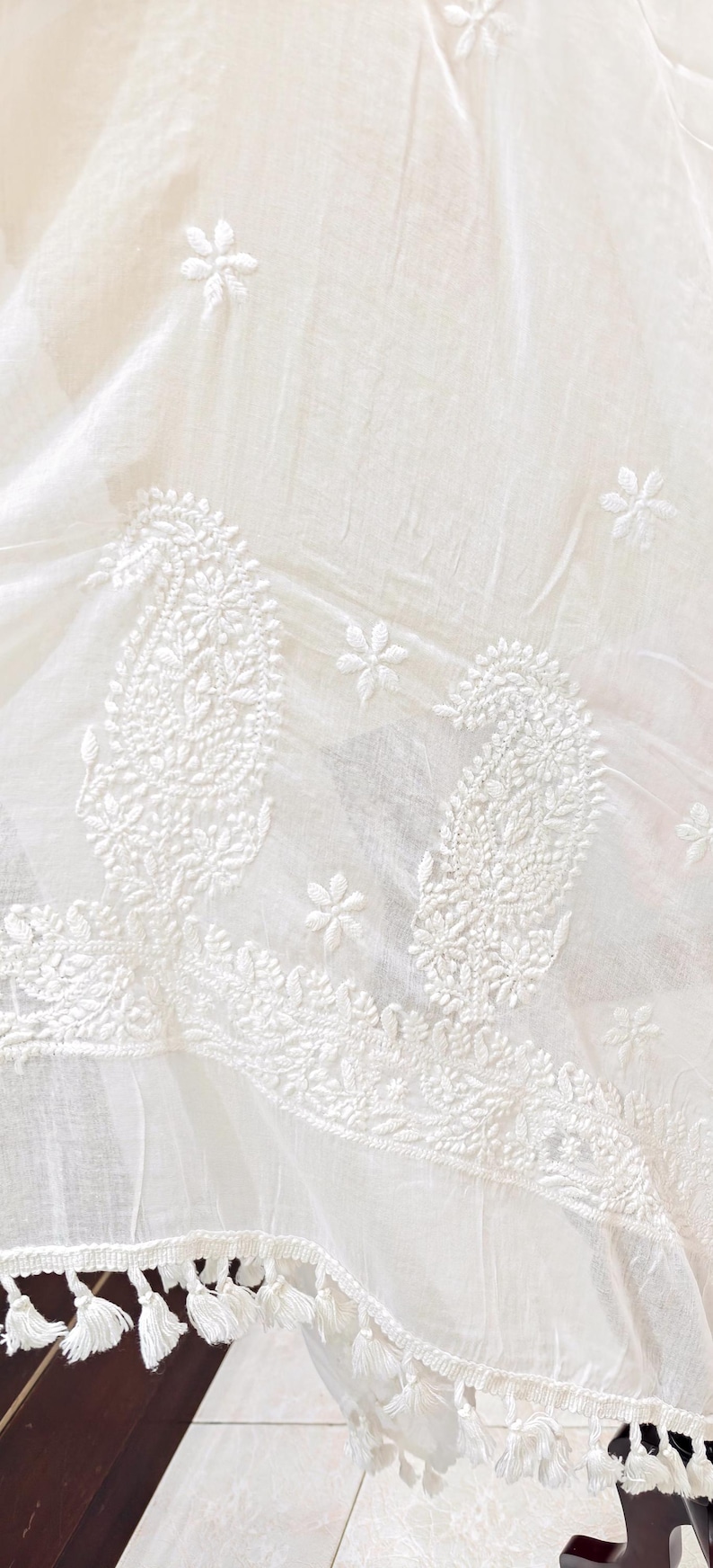 May include: White fabric with intricate embroidered paisley and floral designs. The fabric has a delicate, sheer quality, with a tasseled edge. The embroidery is detailed and adds texture to the textile.