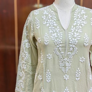 May include: A light green tunic with intricate white floral embroidery. The tunic features a V-neckline and three-quarter sleeves. The embroidery is detailed and covers the front, sleeves, and neckline of the garment.