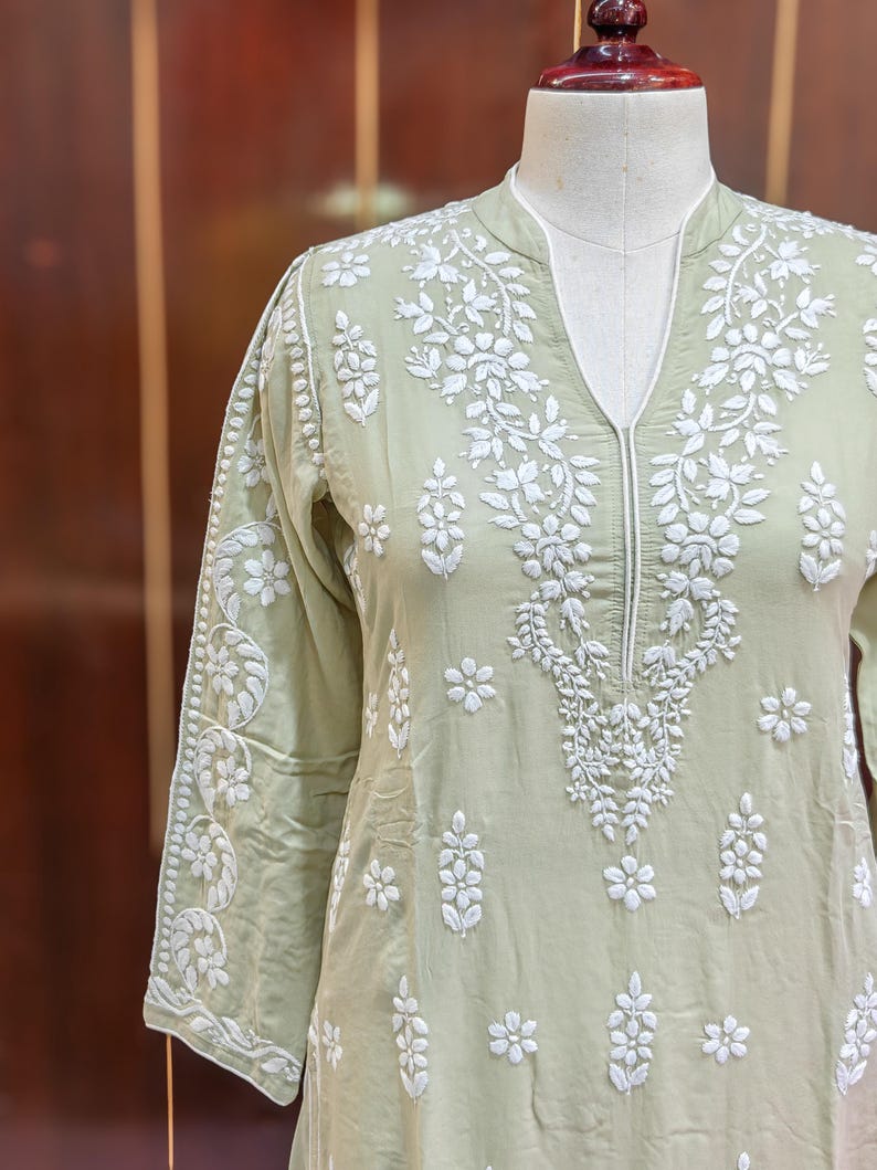 May include: A light green tunic with intricate white floral embroidery. The tunic features a V-neckline and three-quarter sleeves. The embroidery is detailed and covers the front, sleeves, and neckline of the garment.