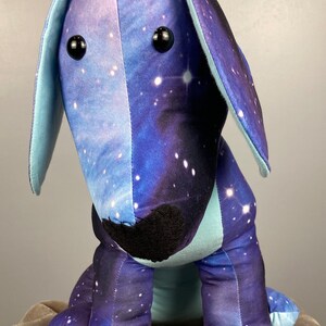 May include: A plush dog toy with a galaxy print in shades of blue, purple, and white. The dog has large, floppy ears, black button eyes, and a black nose. The toy is sitting on a neutral-colored surface.