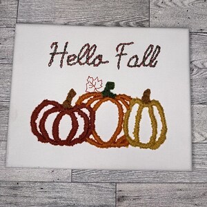 May include: Embroidered white canvas with the text "Hello Fall" and three embroidered pumpkins in shades of orange, yellow, and brown.