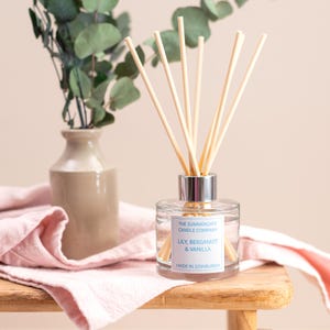 May include: A glass diffuser with wooden reeds, silver collar, and a label stating "Lily, Bergamot & Vanilla". The diffuser is on a wooden surface with a pink cloth and a vase with greenery. Made in Edinburgh.