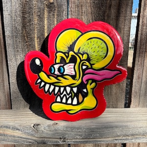 Può includere: Una testa di ratto in stile cartone animato con contorno e sfondo rossi. Il ratto ha pelliccia gialla, lingua rosa e occhi sporgenti. La bocca è aperta, rivelando affilati denti bianchi. Il design è lucido, forse un adesivo o un cartello.