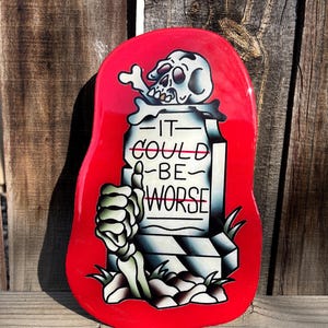 Könnte beinhalten: Ein rotes, tropfenförmiges Dekorationsstück mit einem Design im Tattoo-Stil. Das Design zeigt einen Schädel mit Knochen, einen Grabstein mit dem Text "IT COULD BE WORSE" und eine Skeletthand, die einen Daumen hoch zeigt.