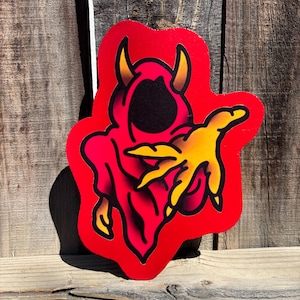 May include: A red, die-cut sticker featuring a devil-like figure in a hooded robe. The figure has yellow horns and a yellow hand reaching out. The design is outlined in black and set against a red background.