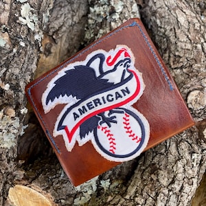 May include: Brown leather wallet with a blue stitched border. The wallet features a patch with a white baseball and a patriotic eagle design with the word "AMERICAN" in red and white.