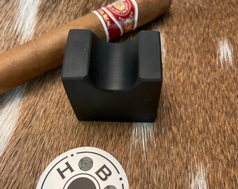 May include: A black cigar rest with a cigar resting on it. The cigar has a red and gold band. A sticker with the text "HOB ORIGINALS" is also present.