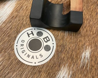 May include: A black cigar rest with a wooden accent, next to a cigar and a circular sticker with the text "ORIGINALS" and the letters "H" and "B". The rest is on a brown and white patterned surface.