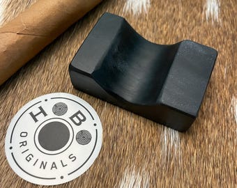 May include: A black cigar rest with a curved design, a cigar, and a sticker with the text "HOB ORIGINALS" on a brown and white patterned surface. The cigar is brown with a red and white band.