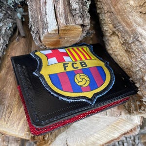 May include: Black leather wallet with a red beaded edge. The wallet features a colorful patch with the FC Barcelona crest, including the team's colors and the letters "FCB". The wallet is set against a backdrop of wooden logs.