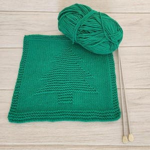 May include: A knitted green square with a Christmas tree design, a ball of green yarn, and two knitting needles. The tree is in the center of the square. The yarn and needles are on the top right of the square.