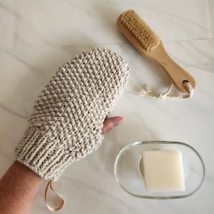 May include: A beige knitted bath mitt, a wooden handled brush with natural bristles, and a bar of soap in a clear glass dish. The mitt has a loop for hanging. The brush has a rope loop. The soap is rectangular.