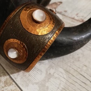 Könnte beinhalten: Ein Kupferarmband mit gehämmerter Textur und zwei weißen Cabochonsteinen, die in der Mitte jedes Kreises eingesetzt sind.