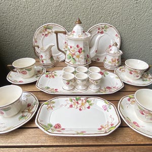Può includere: Un servizio da tè in ceramica con base color crema e motivo floreale di fiori rosa e bianchi con foglie verdi. Il set comprende una teiera, una lattiera, una zuccheriera, quattro tazze con piattini, due vassoi rettangolari e tre portauova.
