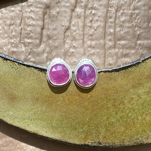 May include: A pair of stud earrings featuring round, faceted, vibrant pink gemstones set in polished silver bezels. The stones display a clear, geometric cut that reflects light, and each earring is secured with a standard metal backing.