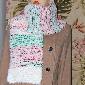 May include: A fluffy, multi-colored scarf with white, red, and green stripes is displayed on a mannequin. The scarf is made of a soft, textured material and is wrapped around the mannequin's neck and chest. The mannequin is wearing a tan coat.