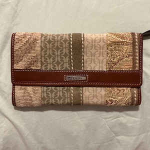 May include: A rectangular Fossil wallet featuring a patchwork design of tan, brown, and beige fabric panels with a repeating logo pattern and textured woven sections. A horizontal brown leather strap with the Fossil logo is centered on the front.