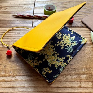 May include: A rectangular wallet featuring a bright yellow flap and a dark blue fabric body patterned with yellow floral clusters. A thin yellow elastic cord with a small red bead charm is attached to the side, securing the folded wallet structure.