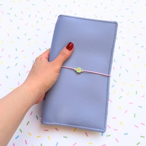 May include: A lilac-coloured journal with a pink elastic band and a small, green and yellow flower charm. The journal is held by a hand with red nail polish. The background is white with colourful confetti.