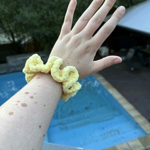May include: A light yellow, handmade scrunchie made of soft, textured fabric. The scrunchie is wrapped around a wrist. A green crocheted item is visible in the foreground. The background includes a pool and trees.