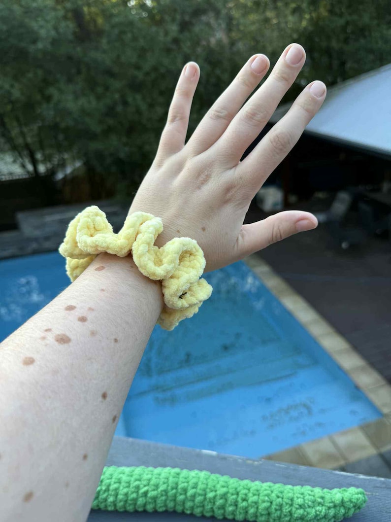 May include: A light yellow, handmade scrunchie made of soft, textured fabric. The scrunchie is wrapped around a wrist. A green crocheted item is visible in the foreground. The background includes a pool and trees.