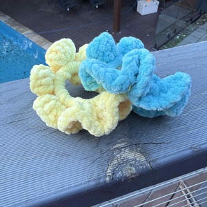May include: Two fluffy scrunchies, one yellow and one blue, rest on a weathered wooden surface. The scrunchies are made of a soft, textured material, perfect for hair accessories. The background shows a pool and outdoor seating.