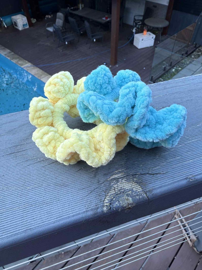 May include: Two fluffy scrunchies, one yellow and one blue, rest on a weathered wooden surface. The scrunchies are made of a soft, textured material, perfect for hair accessories. The background shows a pool and outdoor seating.