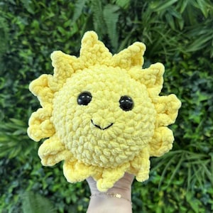 May include: A handmade, yellow crochet sun with a smiling face and black eyes. The sun has a textured, fluffy appearance and is surrounded by spiky rays. The background is a lush green foliage.