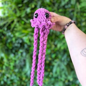 May include: A handmade, plush, purple jellyfish toy with black safety eyes and long, braided tentacles. The jellyfish is held against a backdrop of green foliage. The toy is made of a soft, textured material.