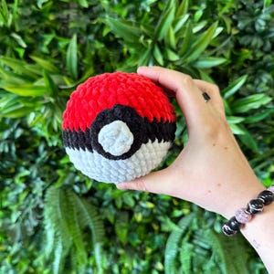 May include: A handmade crochet ball in the style of a Pokeball. The ball is red on top, white on the bottom, and has a black stripe and center circle. The ball is held in a person's hand against a background of green foliage.