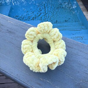 May include: A pale yellow, handmade scrunchie with a textured, looped design. The scrunchie is circular, with a large opening in the center. The scrunchie is made of a soft, plush material. The background is a blue pool and a gray wooden surface.