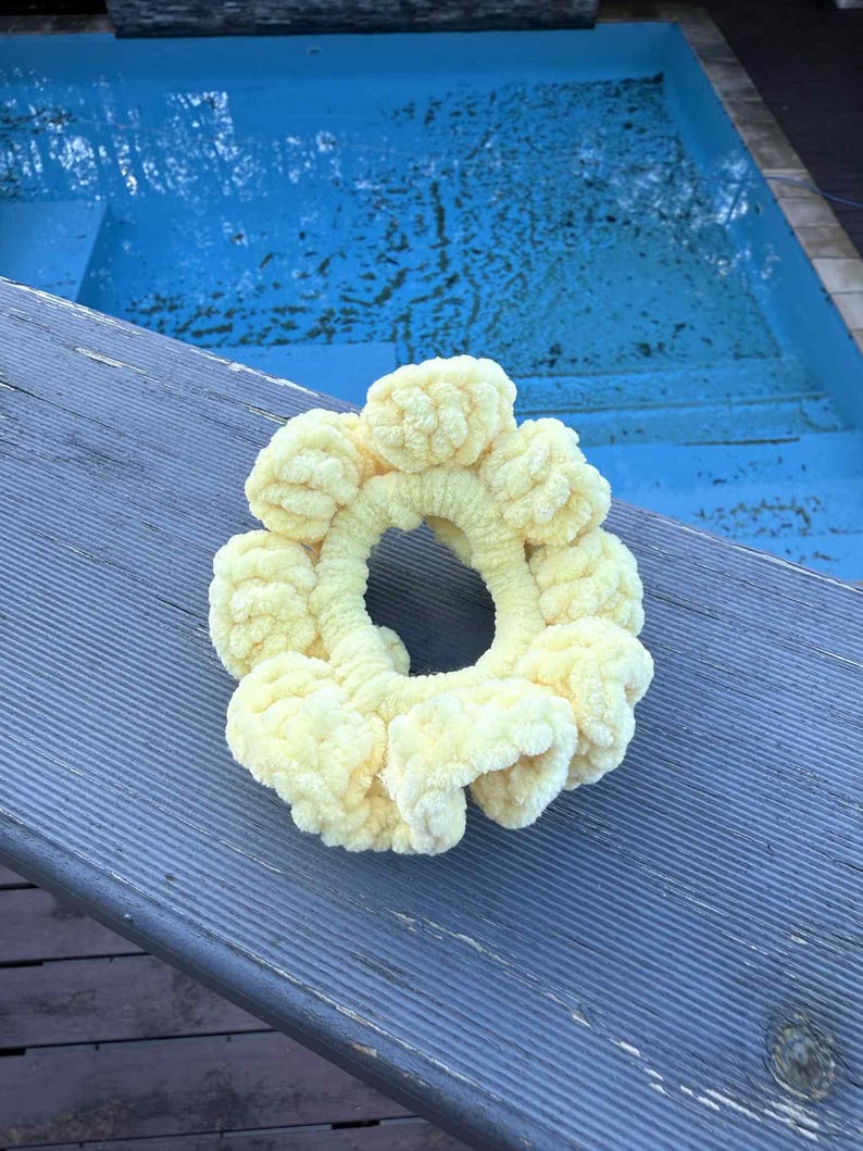 May include: A pale yellow, handmade scrunchie with a textured, looped design. The scrunchie is circular, with a large opening in the center. The scrunchie is made of a soft, plush material. The background is a blue pool and a gray wooden surface.