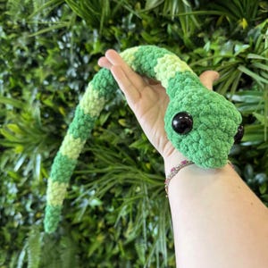 Pode incluir: Um brinquedo de crochê de cobra feito à mão em tons de verde e verde claro. A cobra tem olhos grandes e pretos e é segurada em uma mão contra um fundo de folhagem verde. O corpo da cobra tem aproximadamente 50 cm de comprimento.