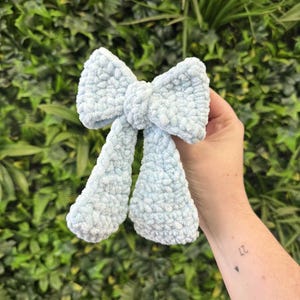 Può includere: Un fiocco all'uncinetto blu chiaro e soffice. Il fiocco è tenuto davanti a uno sfondo di foglie verdi. Il fiocco ha un aspetto strutturato ed è probabilmente realizzato con un filato morbido.