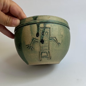 May include: A handcrafted, round ceramic bowl with a green and tan glaze. The bowl features a stylized figure etched into the surface with black dots and lines. The bowl is held by a hand.