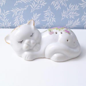 May include: A white ceramic cat figurine in a sleeping pose. The cat has closed eyes and a gold-painted ear. The back of the cat is decorated with small purple and pink floral designs. This decorative item is a salt or pepper shaker.