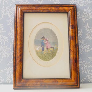 May include: A framed print featuring an oval image of two children kissing in a field of yellow flowers, with a small white dog. The frame is made of dark brown wood with a detailed grain pattern.
